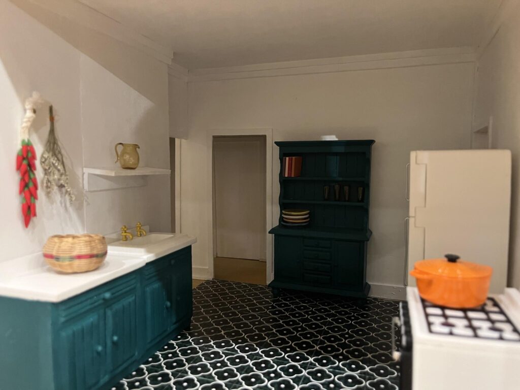 A miniature recreation of a kitchen, with a sink, fridge, and stove, with teal cabinets and checkerboard flooring.