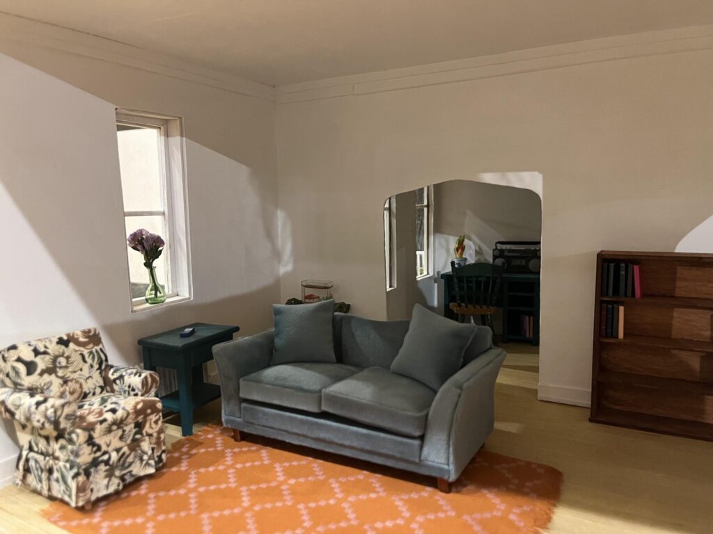 A miniature recreation of a living room, with a blue couch and white patterned armchair, a bookshelf, and a desk.