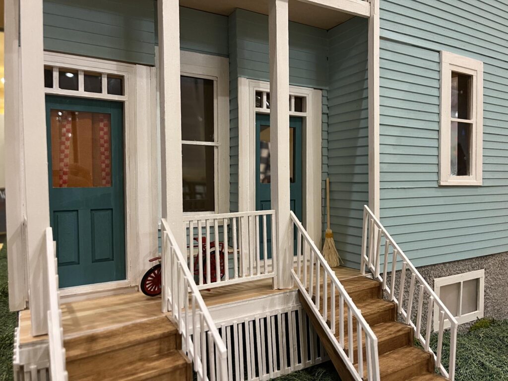 A miniature recreation of the exterior of a house, with blue siding, teal doors, and two sets of steps.