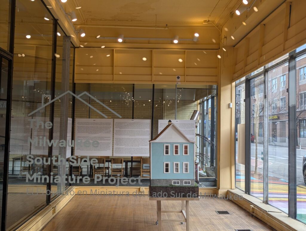 The side of a miniature house in a window display, with signage reading, "The Milwaukee South Side Miniature Project"