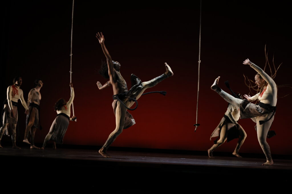 A male and female dancer in the foreground kick the upstage leg toward each other with arms extended away from the body moving up and out. A rope hangs between them. Dancers pose in the background, with one dancer holding on to a rope that descends from above. The dancers wear shades of tan and brown, and red yarn woven to some dancers' tops.