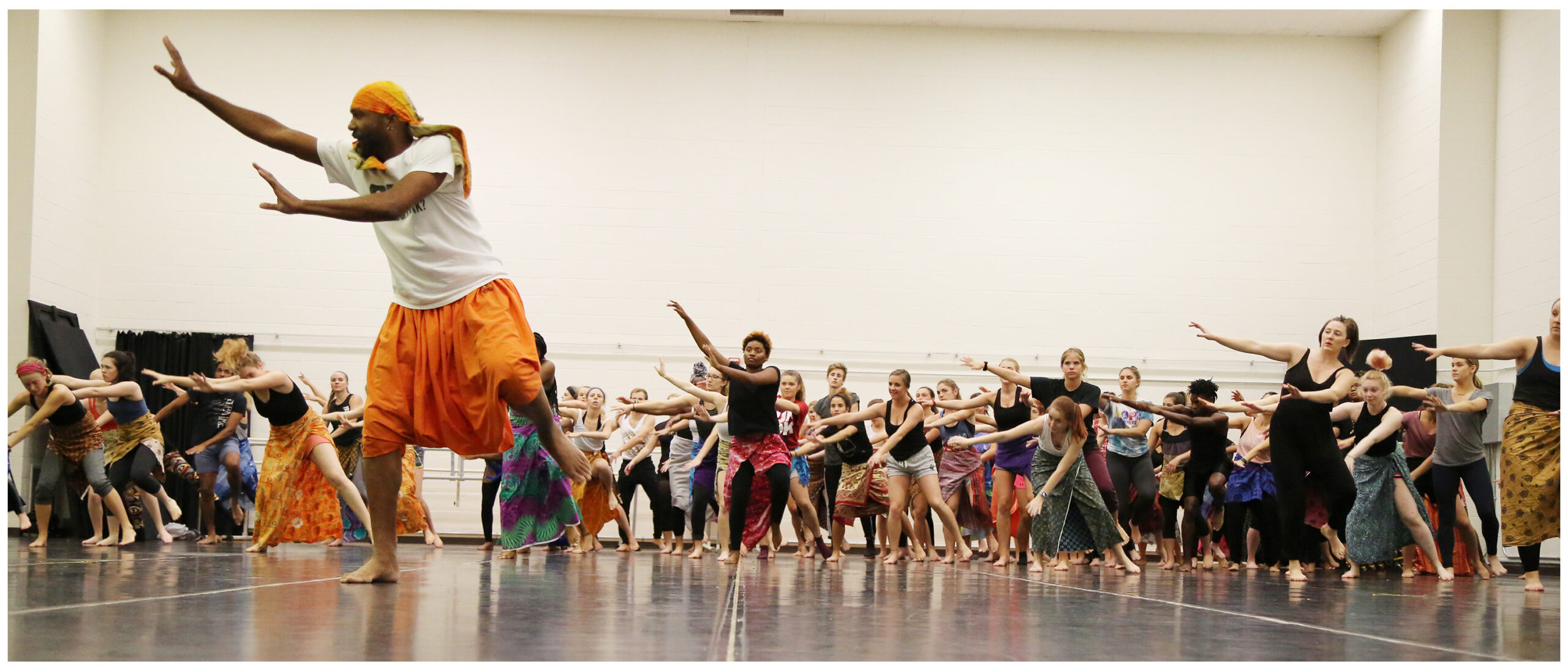 Baba Stafford C. Berry, Jr., wearing orange Chayas (knee-length African pants) and a white t-shirt and orange head tie, demonstrating a dance movement in the foreground, with arms reaching to his right side, while many dancers in the background learn the movement and follow along.