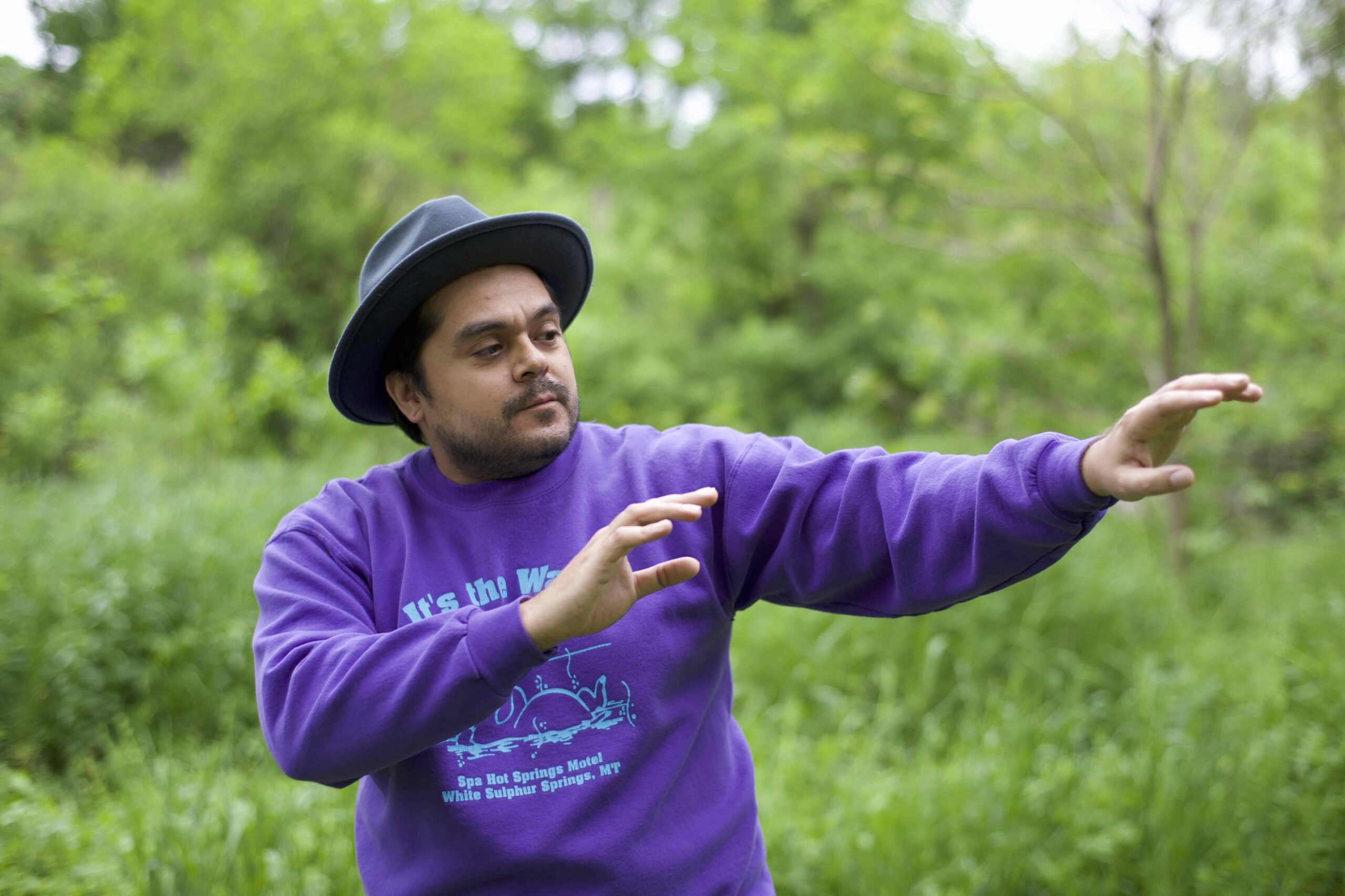 Medium-skin toned man in purple sweatshirt and hat gestures to his left.