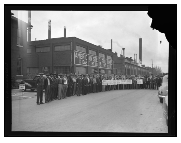 A black and white archival photo of a large group of people standing outside a brick factory building. Some of them are holding placards.