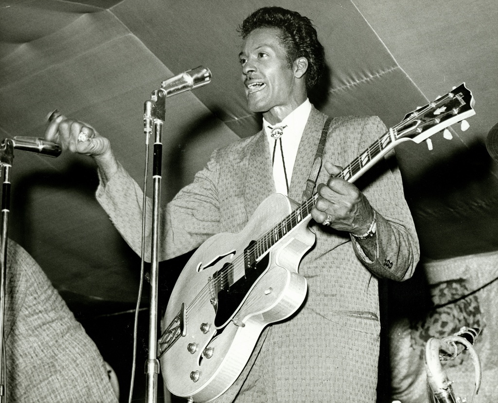 A black and white archival photo of a person singing into a microphone while playing a guitar.