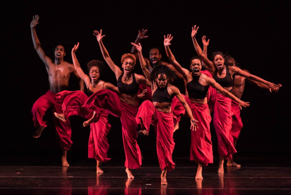 Cluster of dancers reaching right arm and leg upward, with intense facial expressions, wearing red flowing pants and black tops or bare chested.