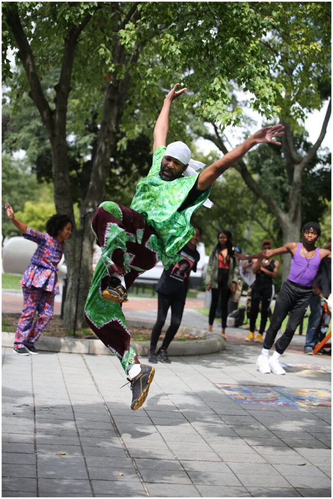 Baba Stafford doing a barrel jump, arms extended over head, wearing green and deep maroon attire, in a park with trees in the background while dancers cheer him on.