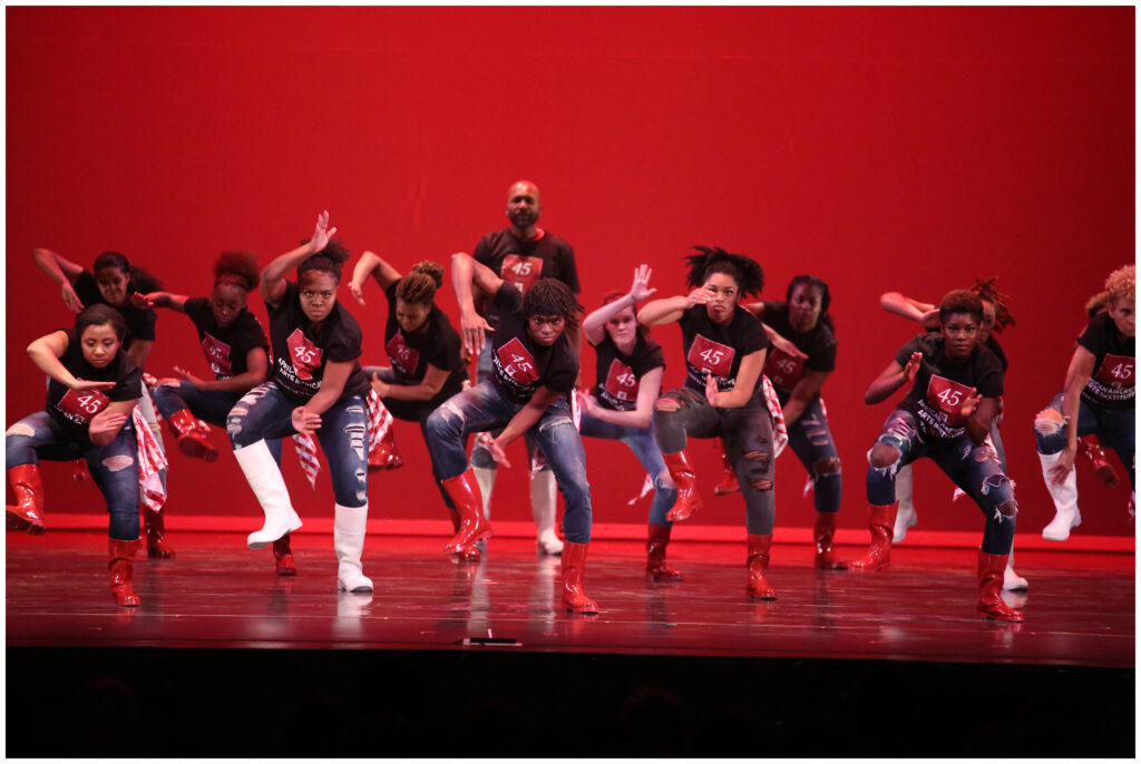 Group of dancers wearing red and white rain boots dancing with arms and feet extended while performing body percussion that includes clapping and stomping. Baba Stafford C. Berry, Jr. stands behind the group of dancers, looking over their heads.