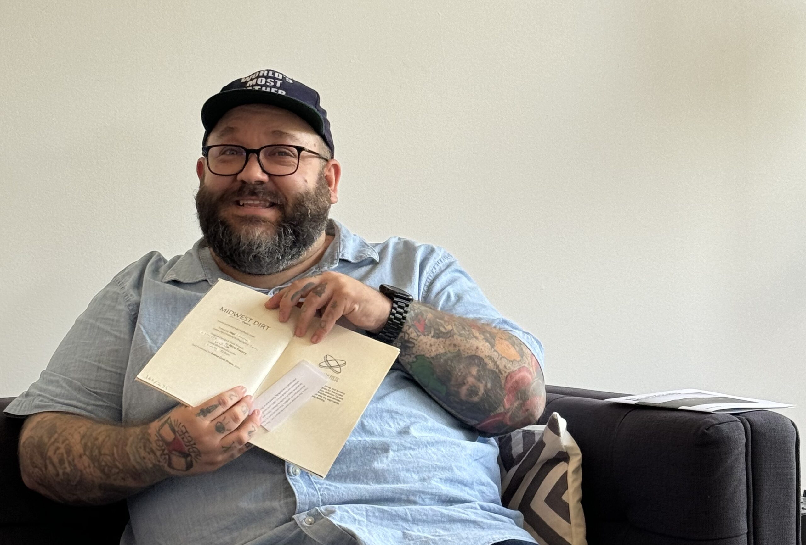 Light-skinned bearded man in baseball cap holds open a zine while sitting on a couch.