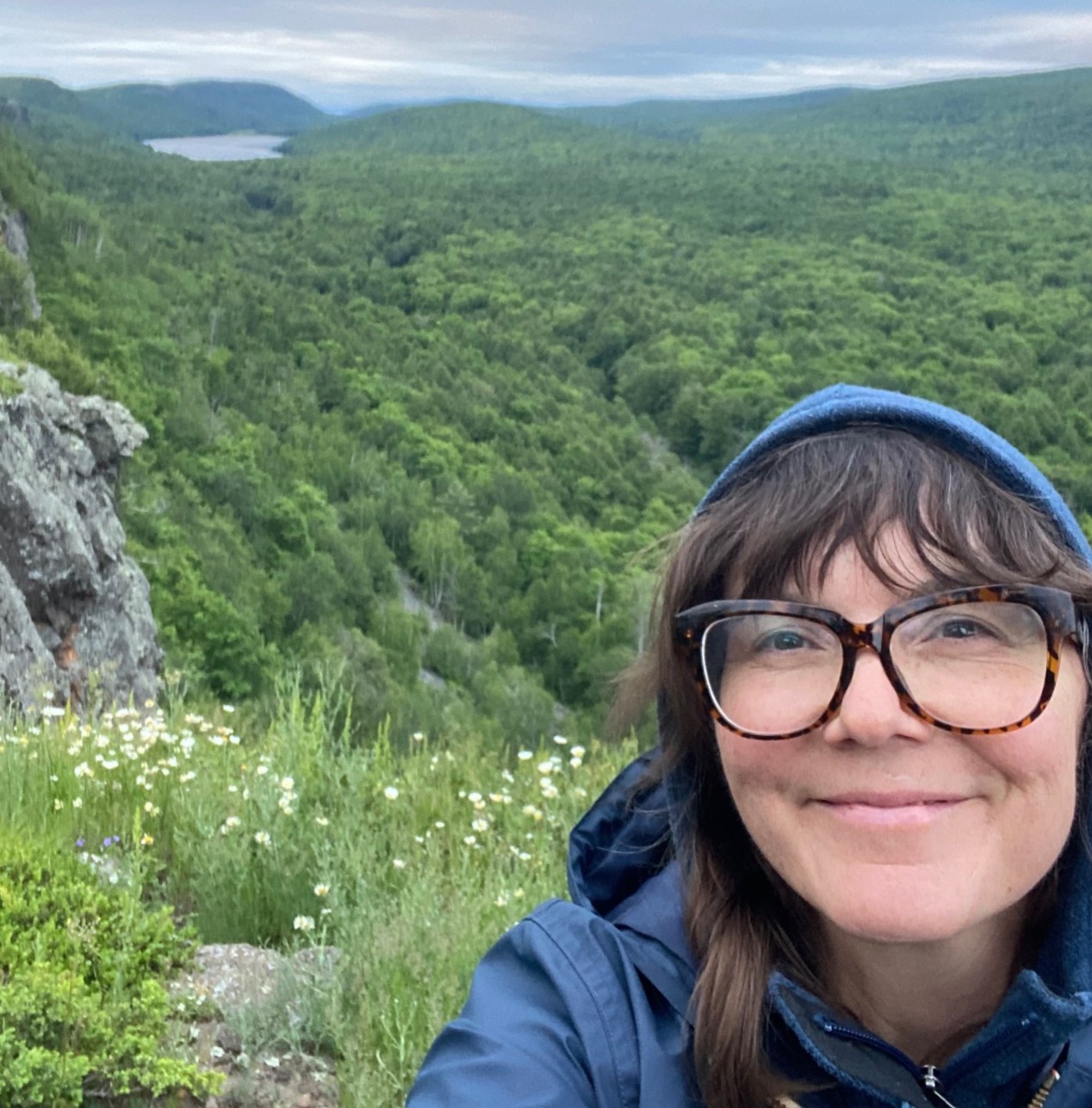 A light-skinned person smiling and wearing glasses and a hoodie while taking a selfie in front of trees on a vista.