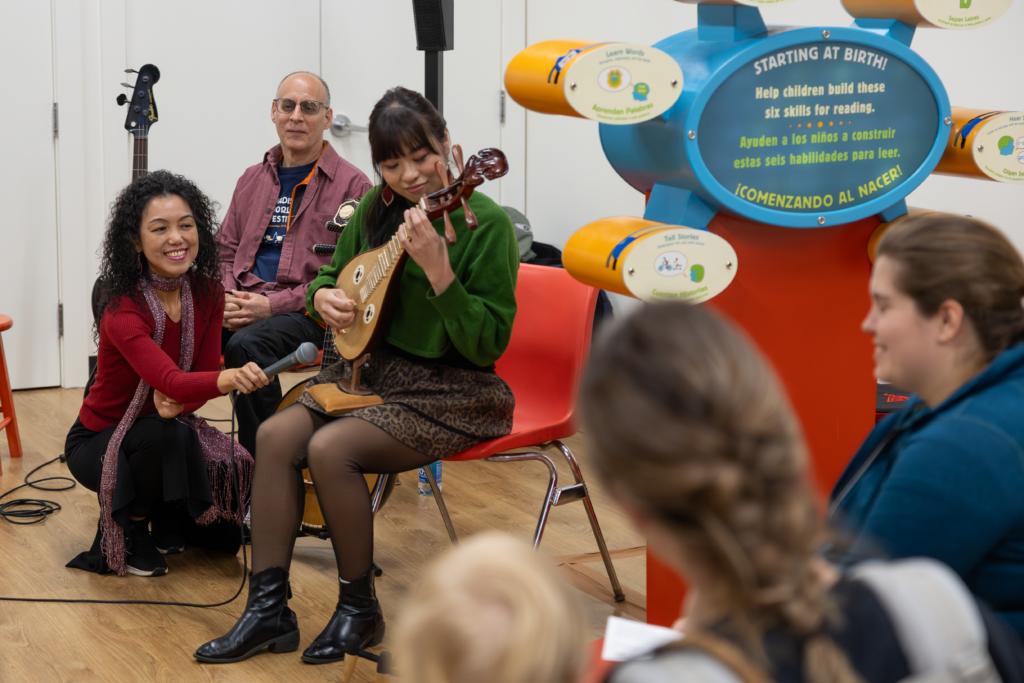 A woman plays a small string instrument as another woman holds a microphone to it.