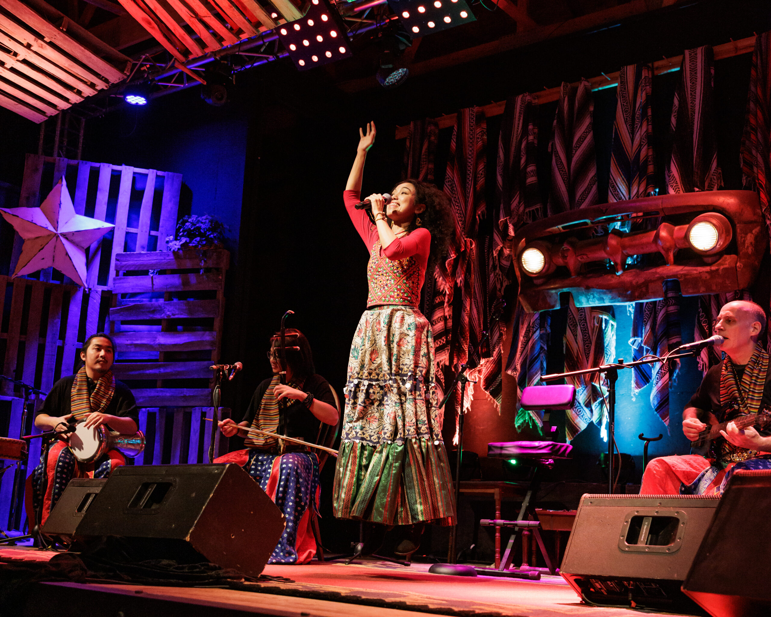 A woman holds a microphone in one hand and has her other arm raised as she jumps in the air, with three band members sitting onstage behind her.