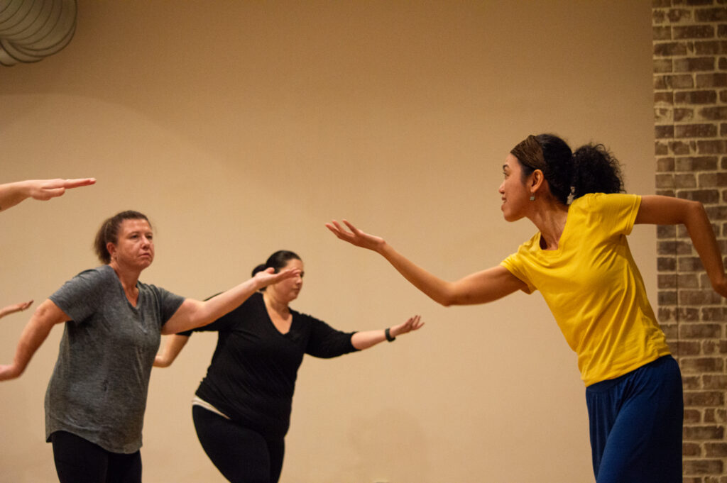 A woman in a yellow top leans forward with an outstretched arm as two others facing her mimic her pose.