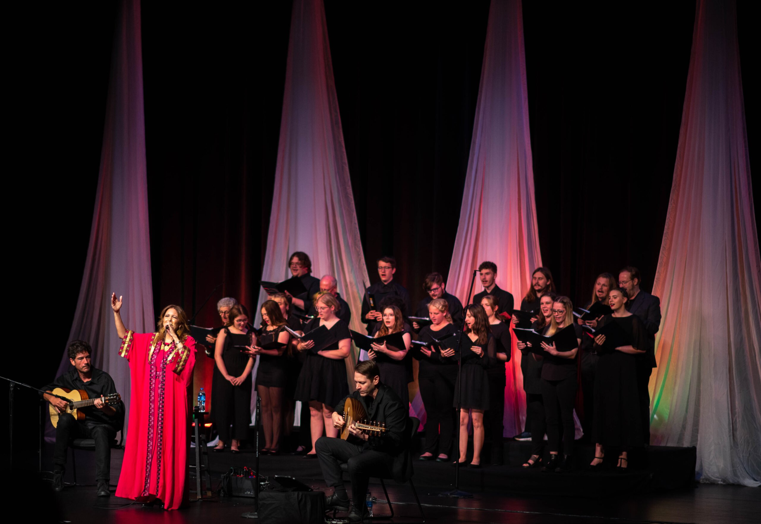 A woman in a long pink dress holds a microphone and sings as a group of 15 people wearing black and holding black folders sing behind her.