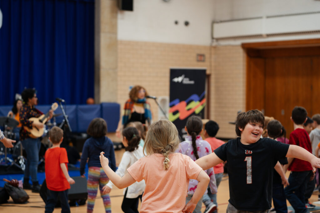 A boy and other students dance and smile in the foreground as musicians perform in the background.