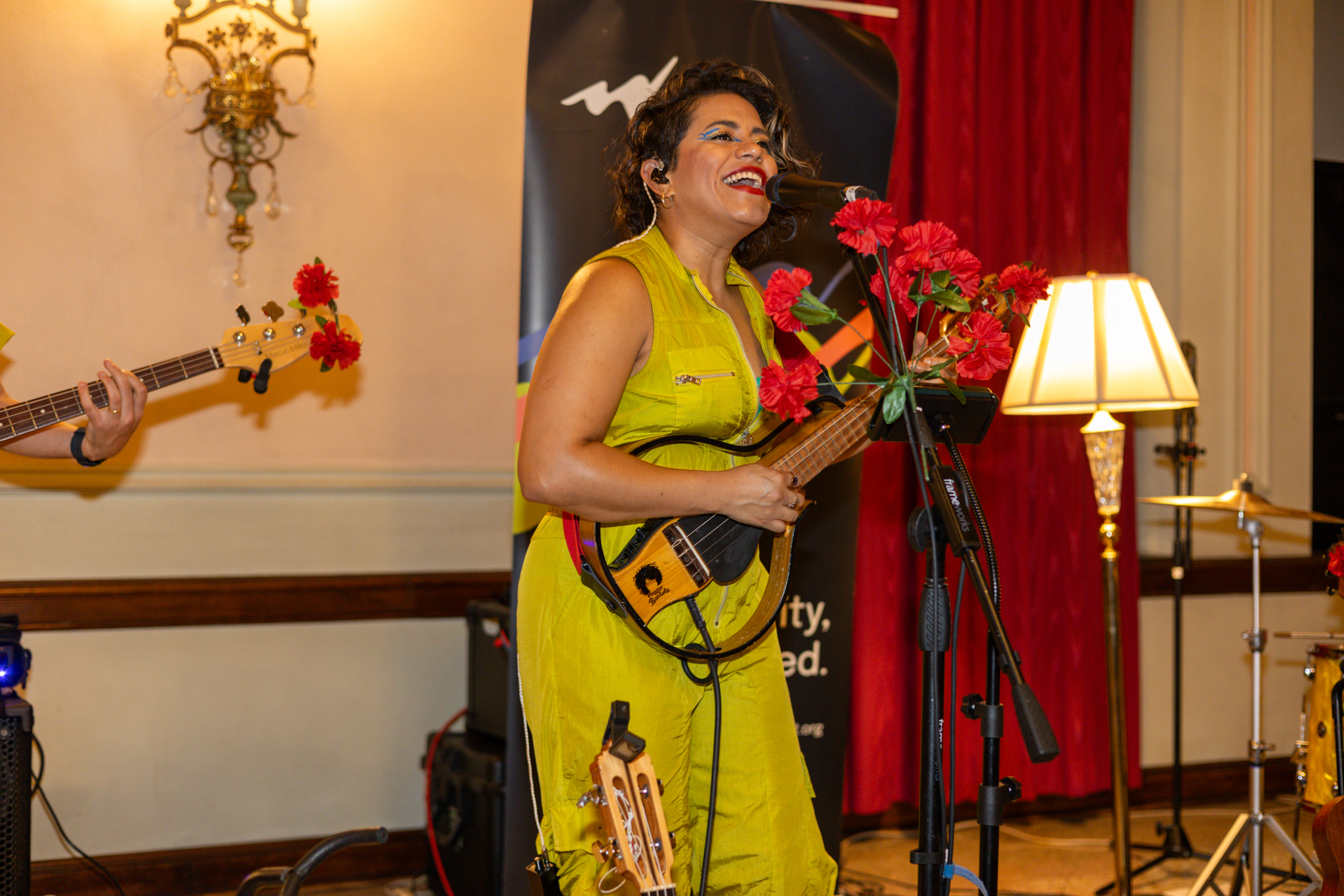 A woman wearing neon yellow sings into a microphone and plays an electronic string instrument.