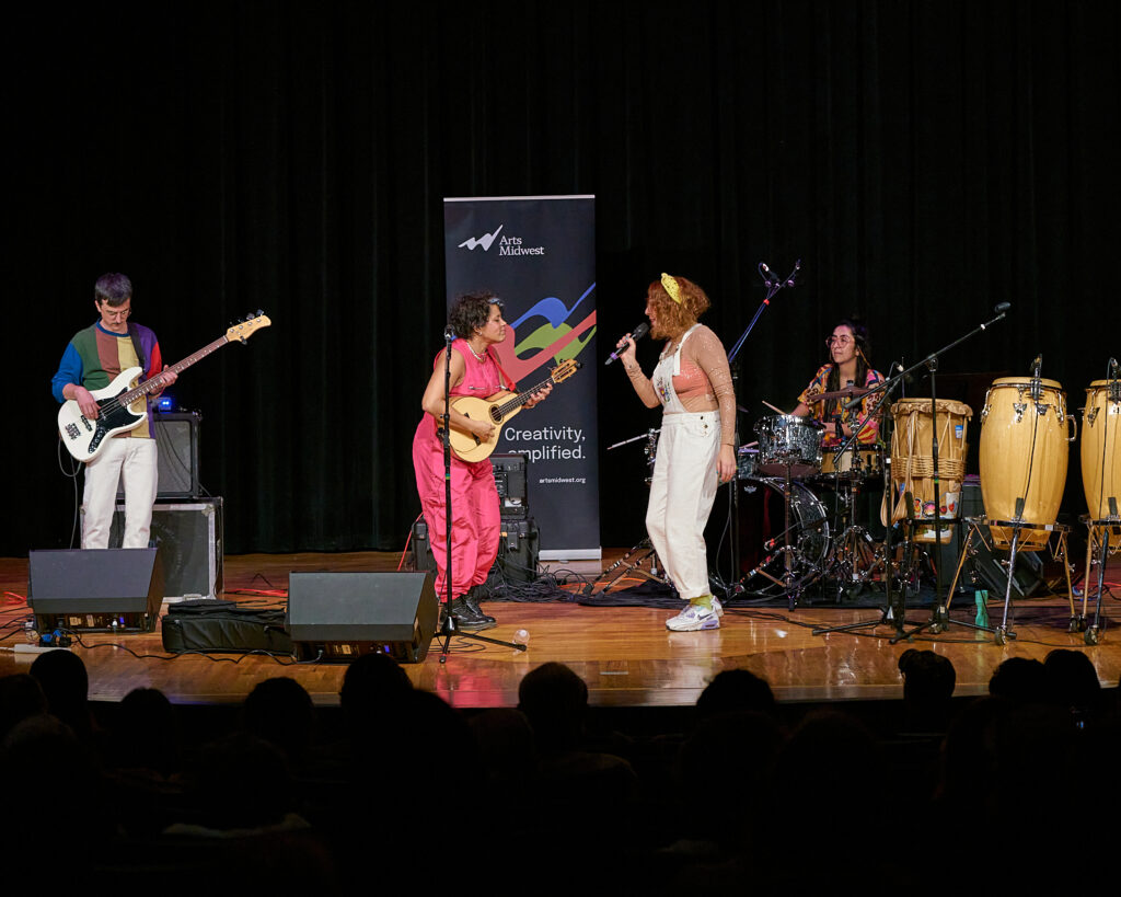 Four people sing and play instruments on the stage of an auditorium, with the heads of audience members in the foreground.