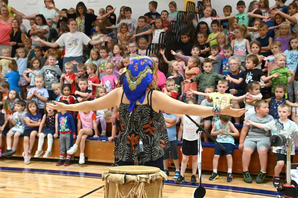 A woman with outstretched arms is seen from behind, facing a large number of middle school aged students on bleachers who are mimicking her pose.