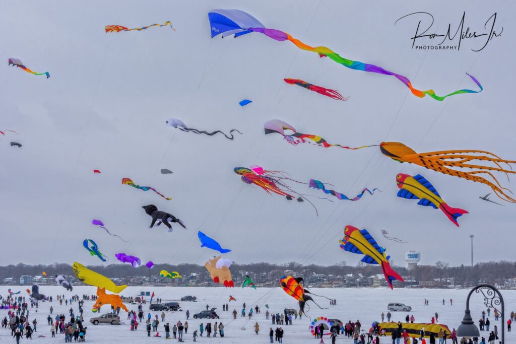 Large colorful kites fly across a frozen lake against a white sky.