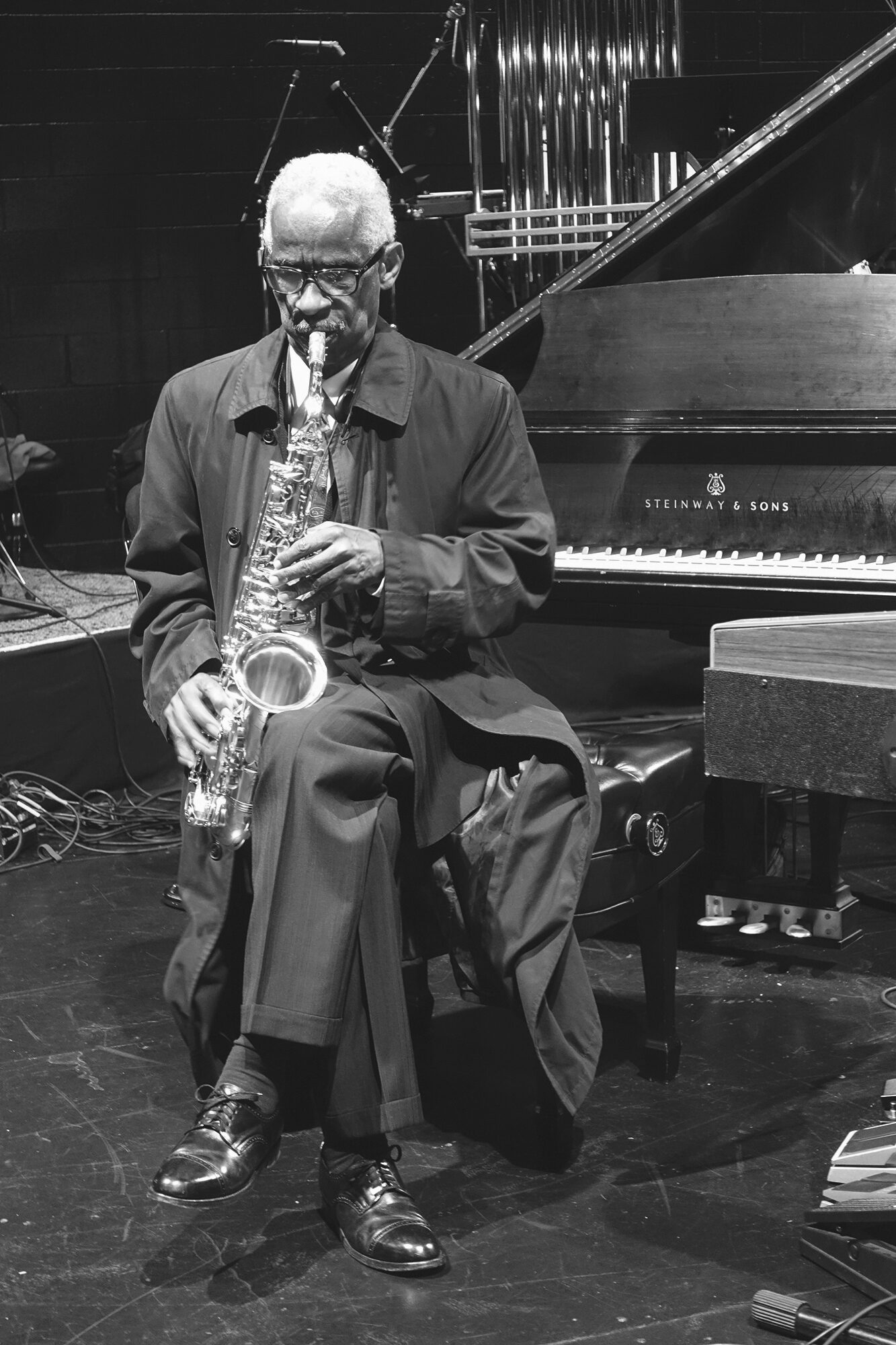 An elderly person with short white hair in a long overcoat sitting and playing a saxophone.