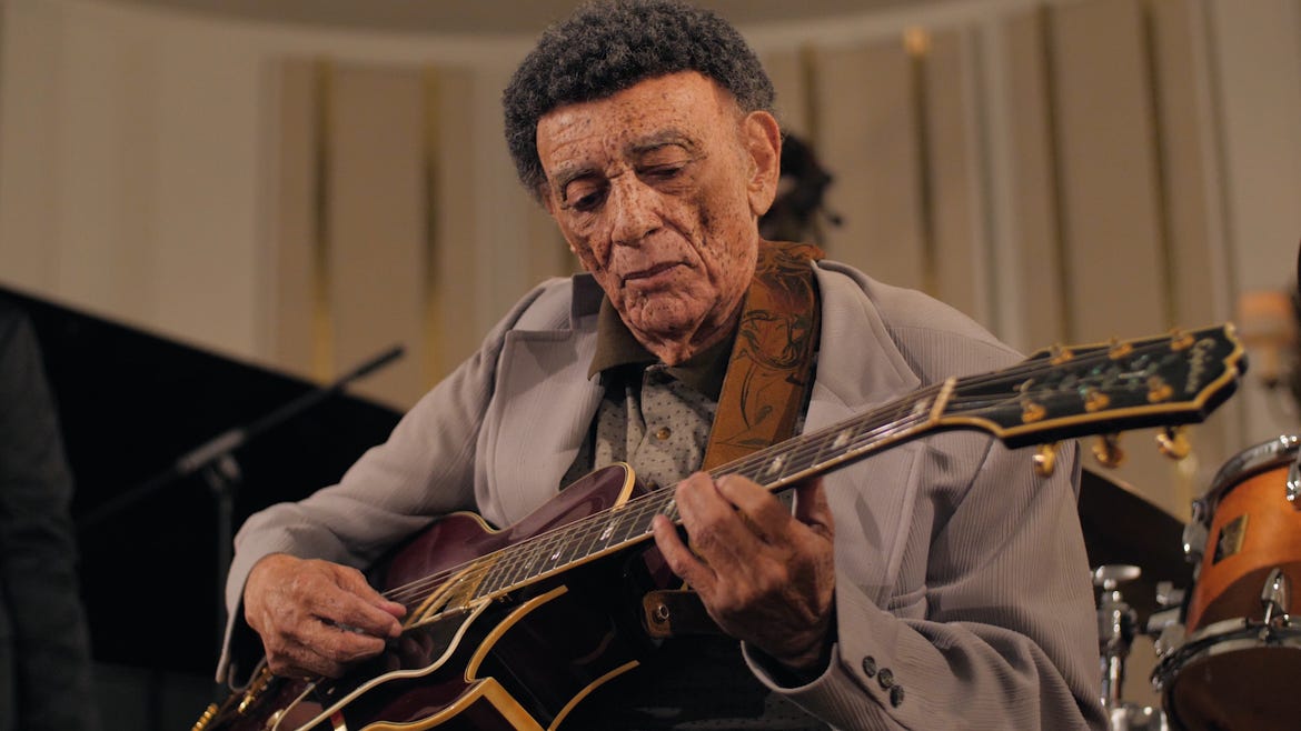 An elderly person with dark hair wearing a grey jacket playing a guitar.