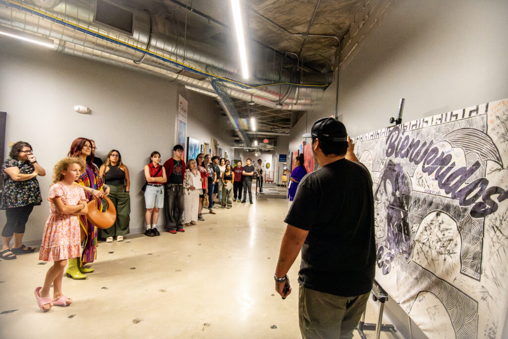 A group of Latinx spectators of different ages listen and watch attentively as the artist points to a large print that says Bienvenidos A Des Moines (Welcome to Des Moines).