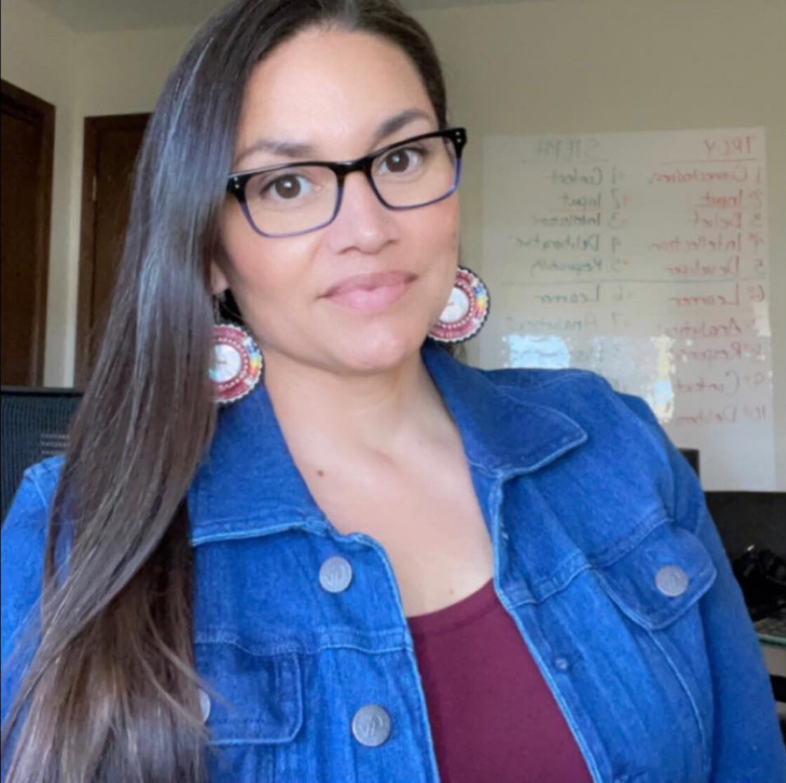 A person of medium skin tone wearing glasses, beaded earrings, a dark red top with a blue denim jacket over it. They have long dark hair over one side of their shoulder.