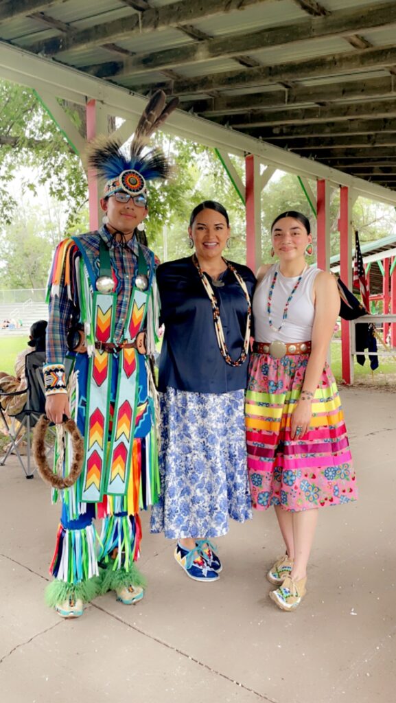 An adult with two young adults standing under an outdoor structure. One of the young adults is wearing traditional regalia, while others have accessories.