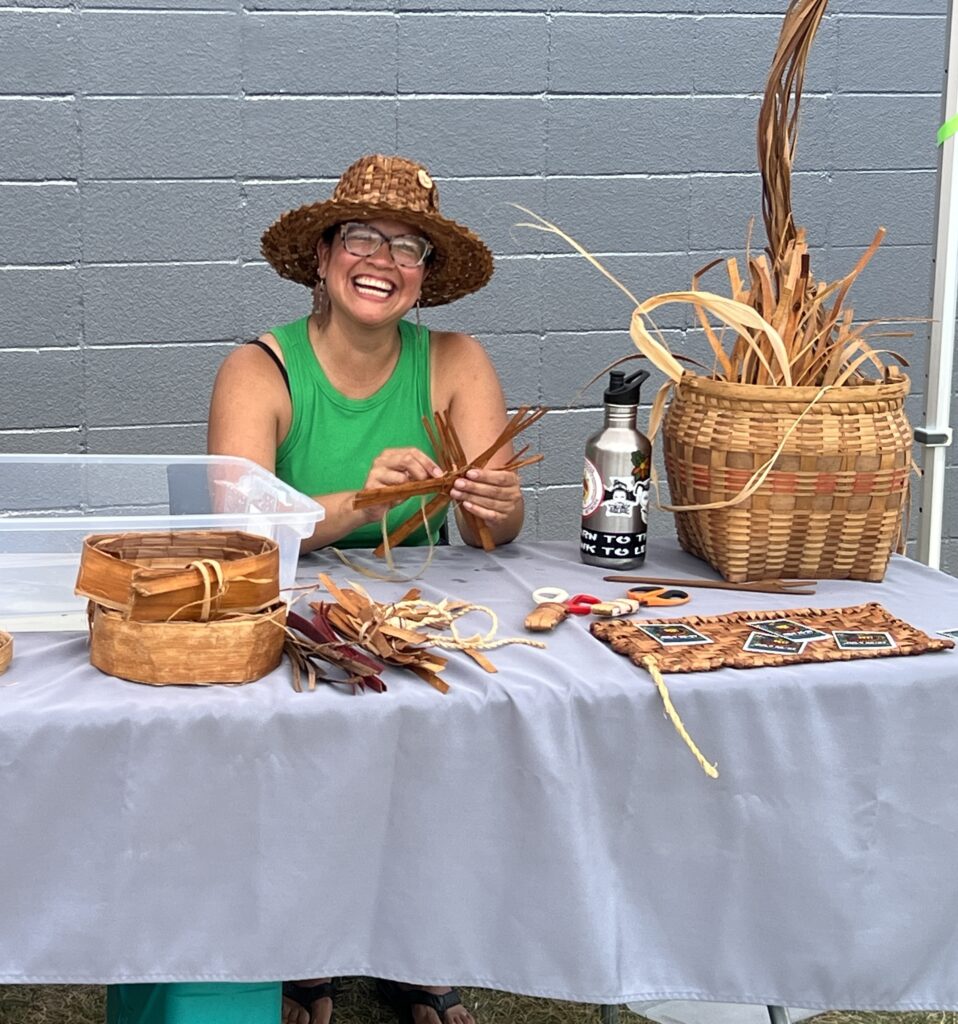 A person wearing a hat sitting at a table with Ojibwe traditional baskets and weaving materials.