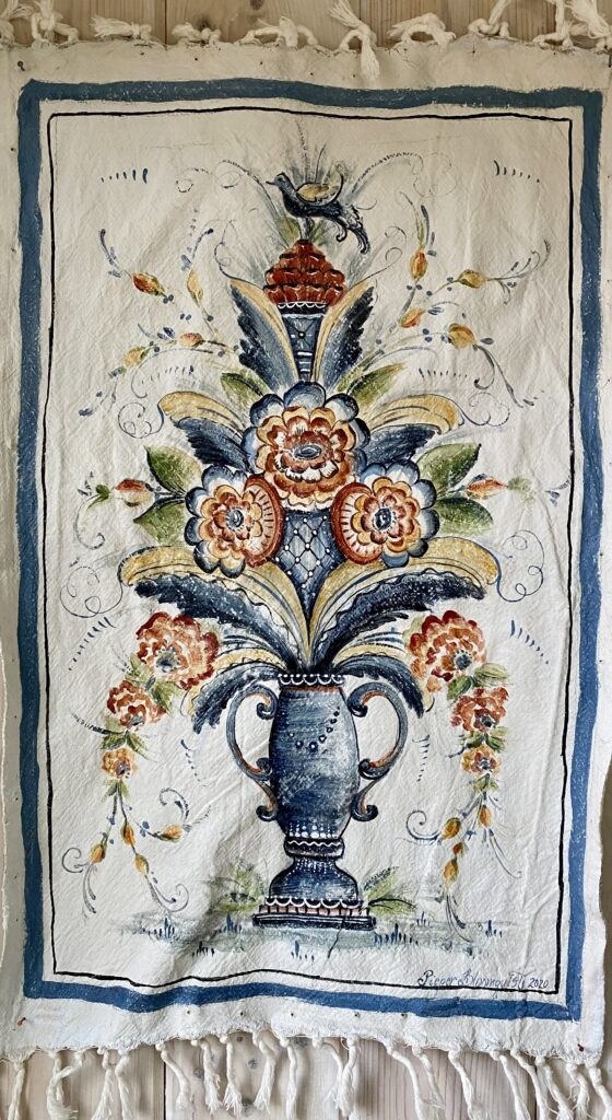 A tall narrow white painted cloth with tasseled top and bottom edges, featuring a painting of a vase with flowers and a bird.