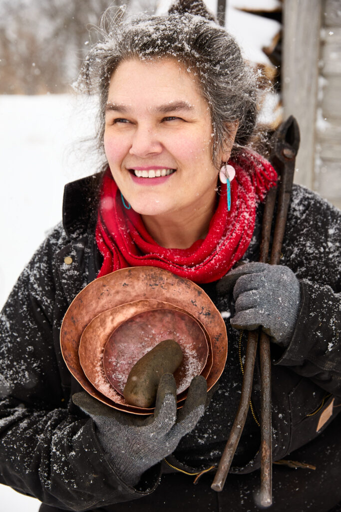 A person of medium-light skin tone and silver hair in the snow, holding several copper bowls.