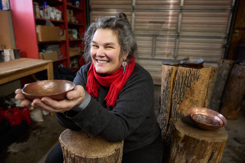 A person of light skin tone and silver hair smiling and holding a copper bowl.