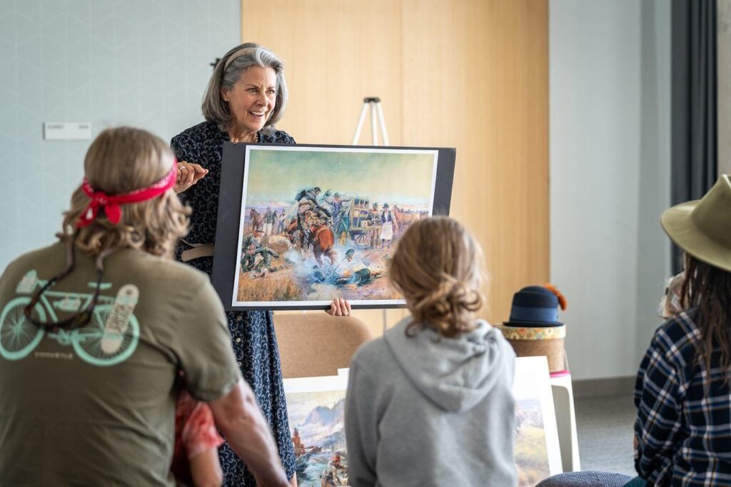 An enthusiastic woman with grey hair holds a painting depicting a wild west scene in front of a group of families.