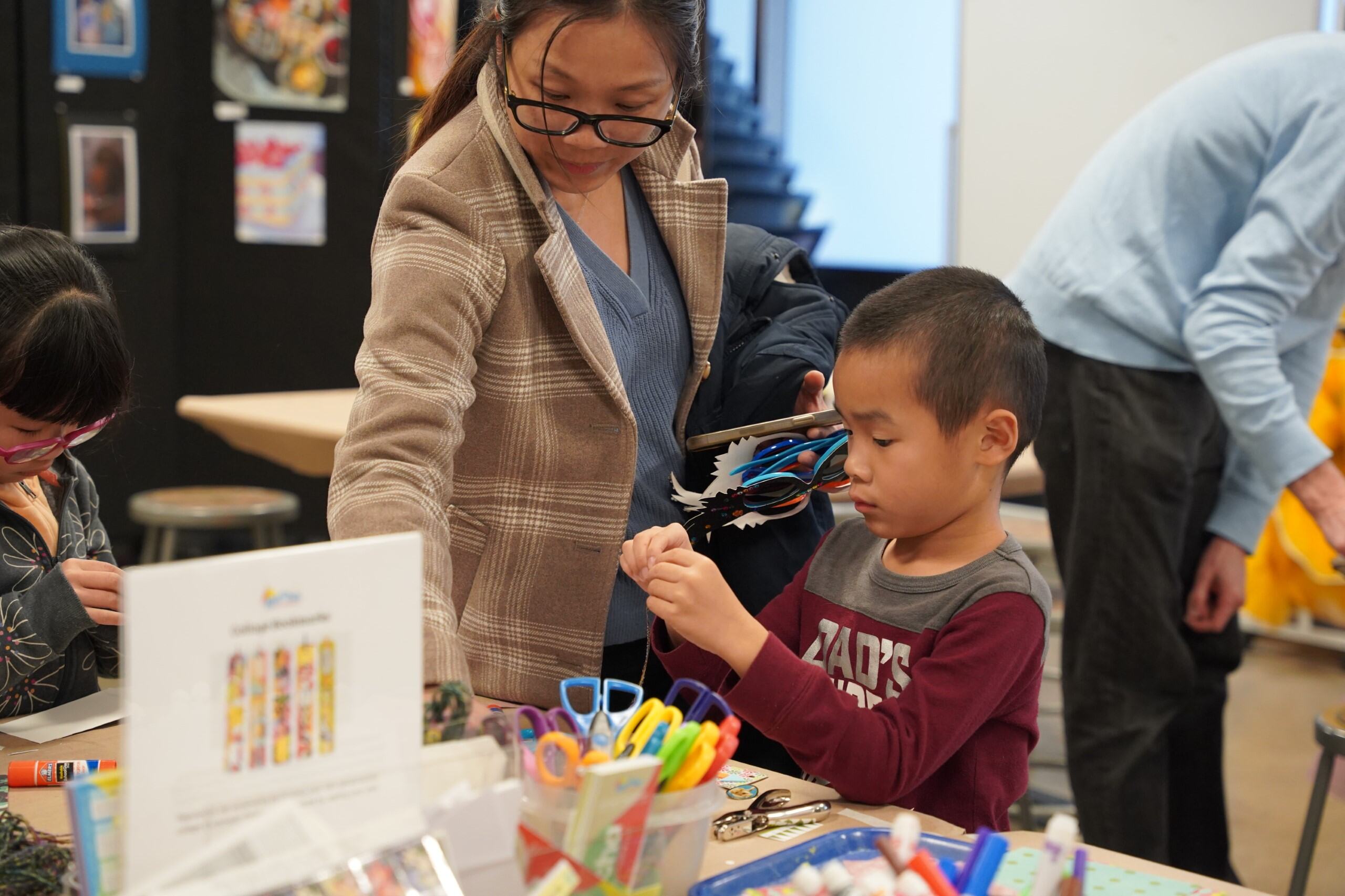 A woman wearing glasses instructs a young boy at an arts and crafts table.