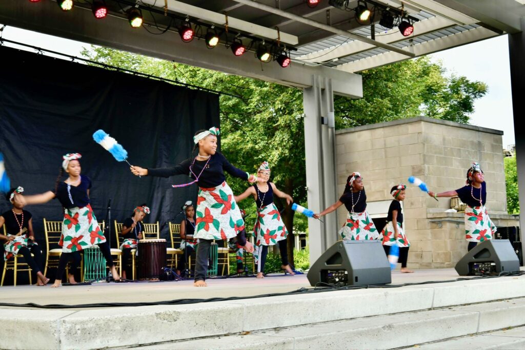 A group of young dancers from Bi-Okoto's Okoto Kekere (Pre-professional group) performing outdoors at Washington Park, during the World Drum Festival in Cincinnati, Ohio, on July 21, 2024. The children are dressed in colorful outfits with floral-patterned skirts, black tops, and matching headwraps. Each holds a blue and white fly whisk, used as a traditional African dance prop. They are mid-movement, showcasing synchronized, rhythmic dance steps. The performance takes place on a stage with a black backdrop and trees in the background, while drummers seated behind them play traditional African drums.