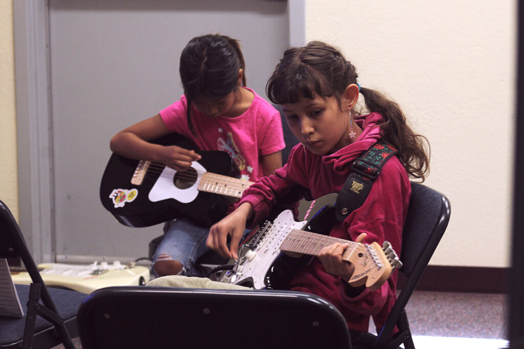 Two youth with dark hair wearing pink shirts sit in black folding chairs, the child on the left playing a black and white mini acoustic guitar, and the youth on the right is playing a black and white electric guitar. Both are peering down at their instruments as they learn the chords.