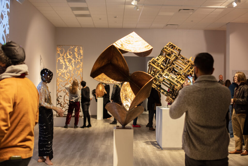 Several audience members and a performer wearing a mask and decorative garb mingling among metal sculptures in the exhibition gallery