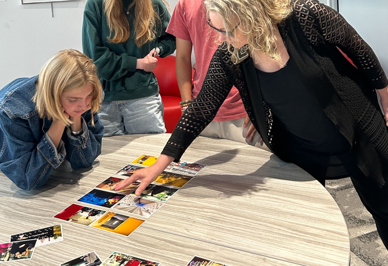 White-skinned person leans over a table and points at pictures while another white-skinned person looks on.