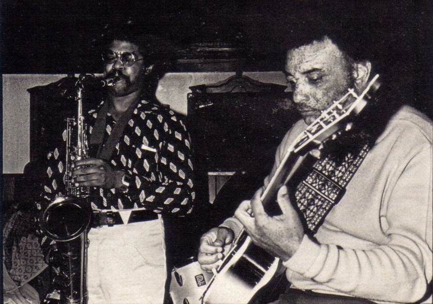 An archival black and white photo of two musicians. One playing a saxophone and the other a guitar.