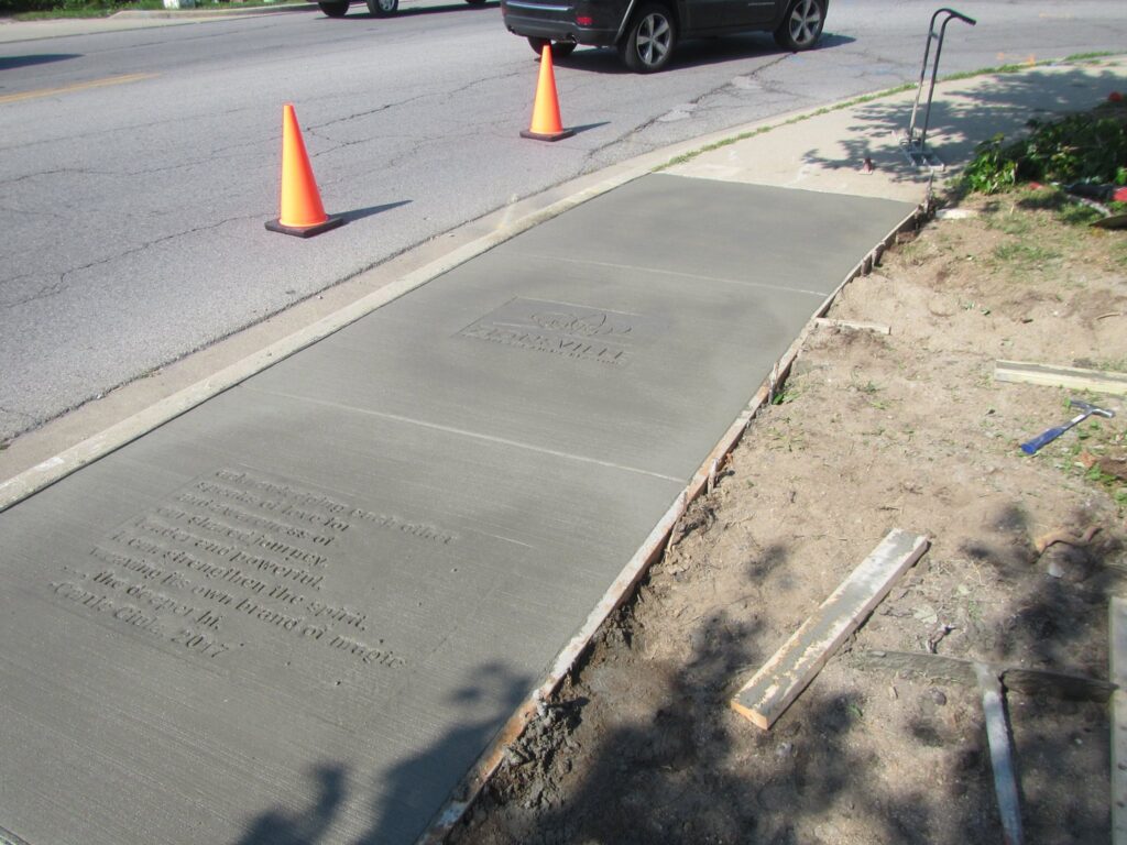 A wet-concrete sidewalk with poems stamped into it next to orange traffic cones.