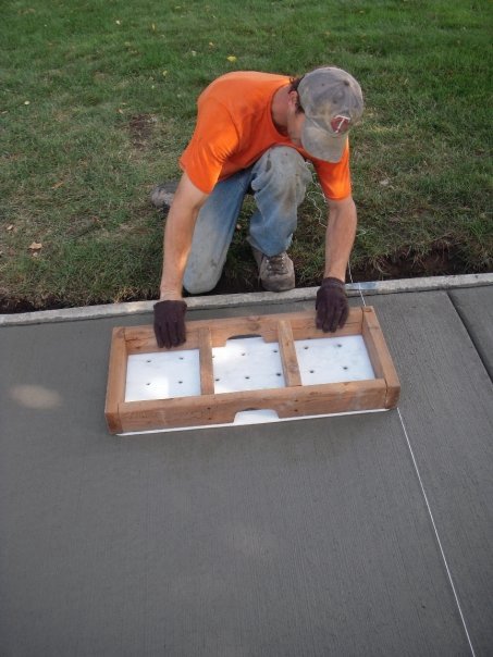 A light-skinned person in a baseball cap, orange shirt, and gloves lies a large stamp framed with wood on a sidewalk next to green grass.