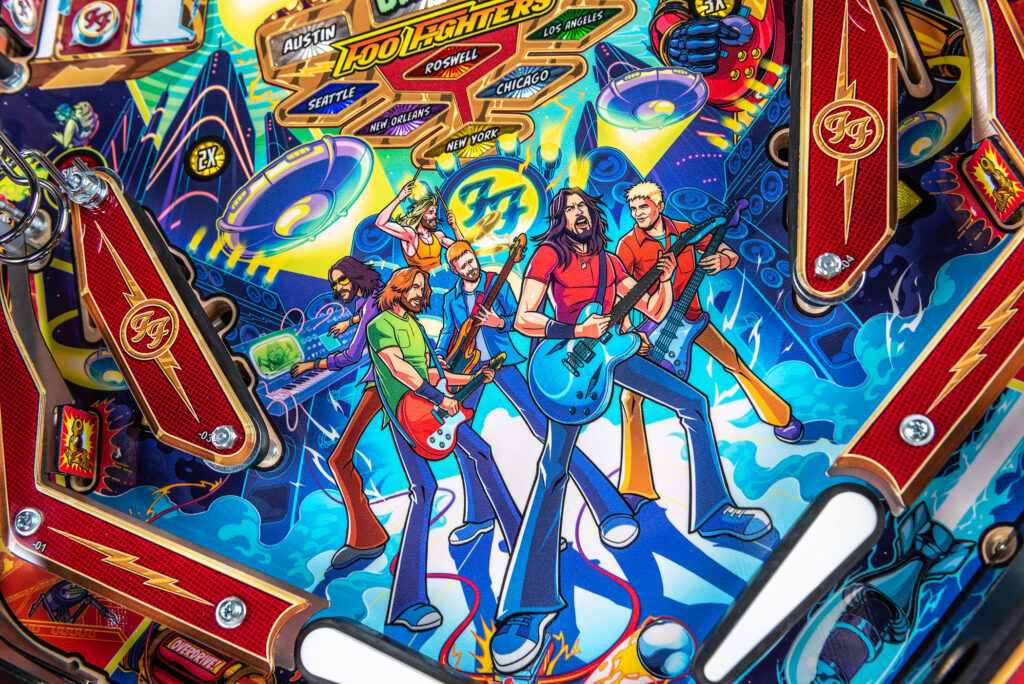 Playfield artwork of the Foo Fighters pinball machine featuring cartoon illustrations of the band members performing on stage, surrounded by speakers, flying saucers, and city signposts like Roswell and Chicago.