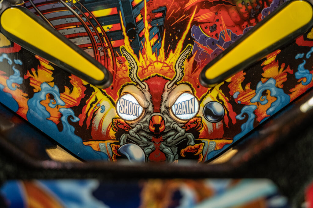 Close-up of the “Shoot Again” insert on the Godzilla pinball machine, illustrated with a fiery-eyed insect creature with pinball eyes in a comic book explosion style.