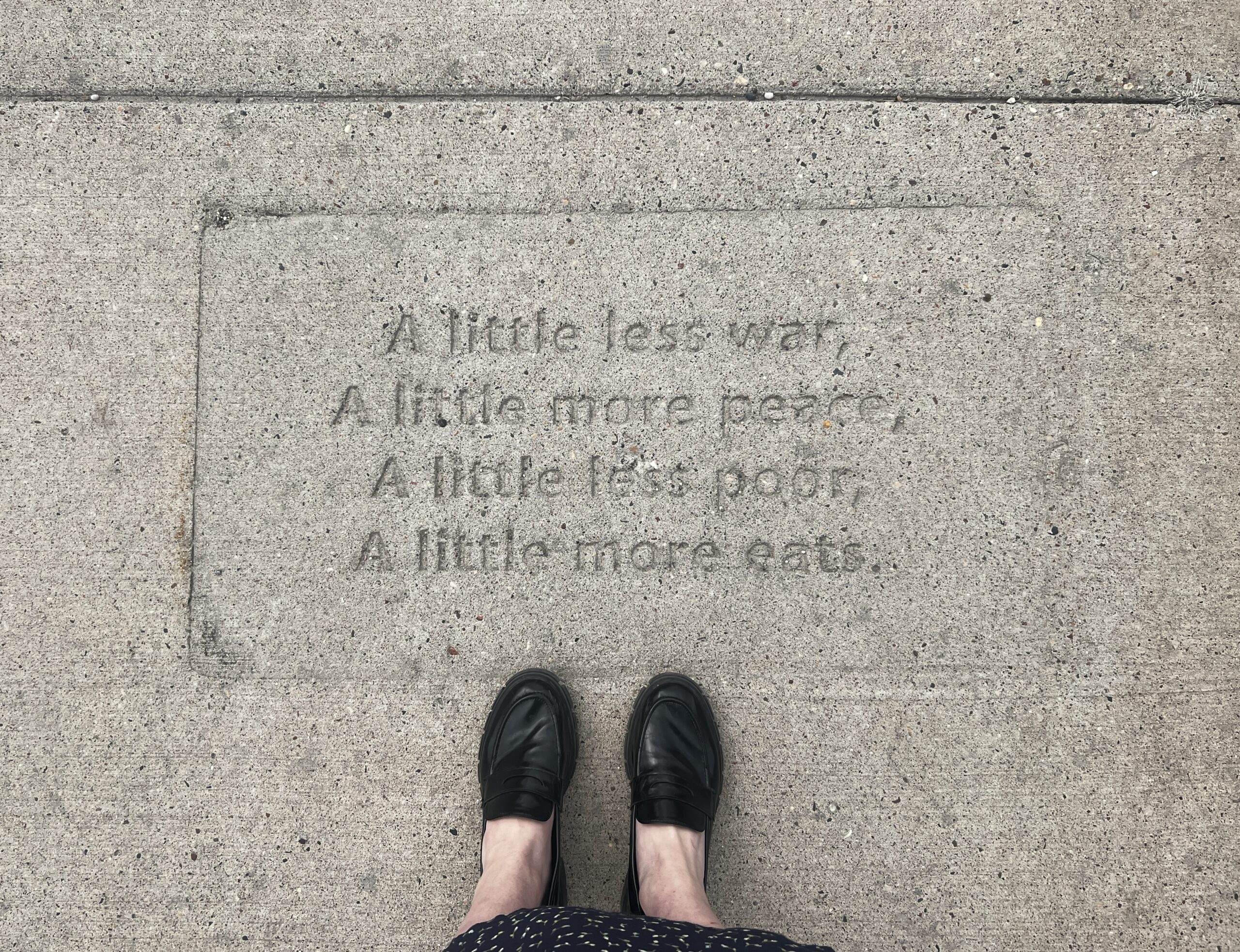 Two feet stand next to a poem stamped into a sidewalk, reading: "A little less war, a little more peace, a little less poor, a little more eats."