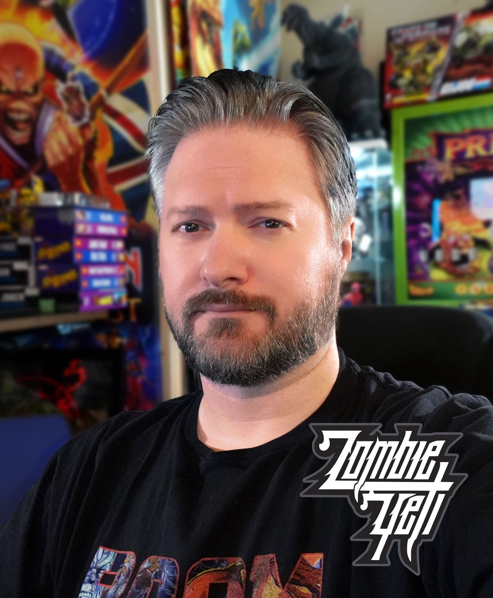 A headshot of a man with light skin and greying hair and a beard, posing in front of a chaotic background of pinball machines