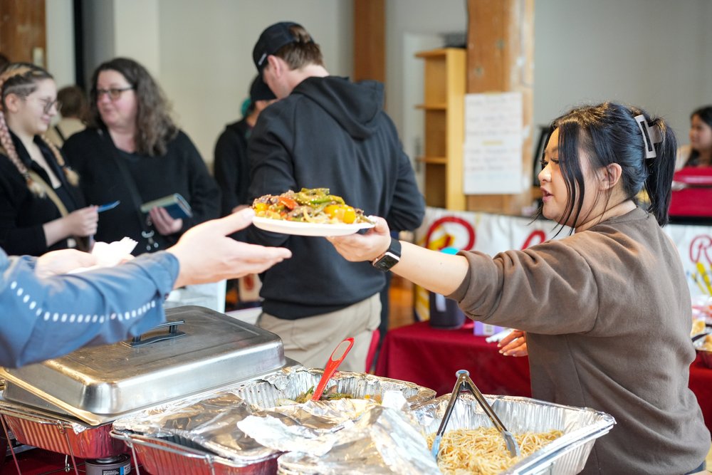 A medium-skin-toned person hands over a dish full of food to a hand outstretched across a table.