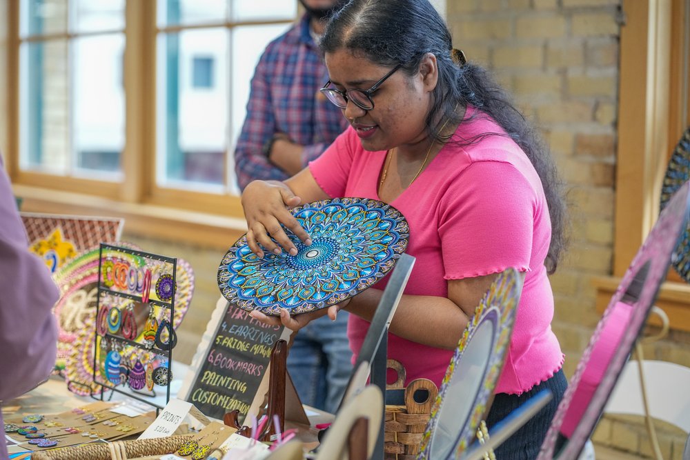 A medium-skin-toned person wearing a bright pink shirt stands and holds a blue, circular art piece above a table.