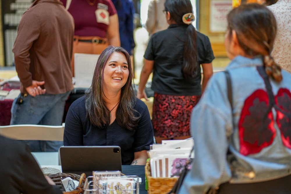 A person with long dark hair smiles and sits behind a table while facing a customer.
