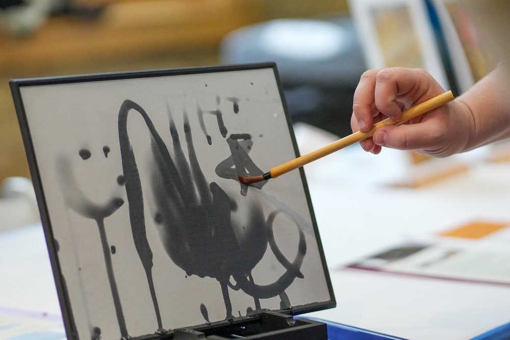 A hand holds a paintbrush that draws dark, ink-like shapes on a white board.