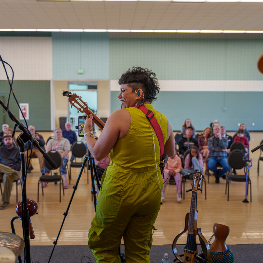 A guitarist in a bright yellow jumpsuit smiles while performing for a seated audience in a multipurpose room.
